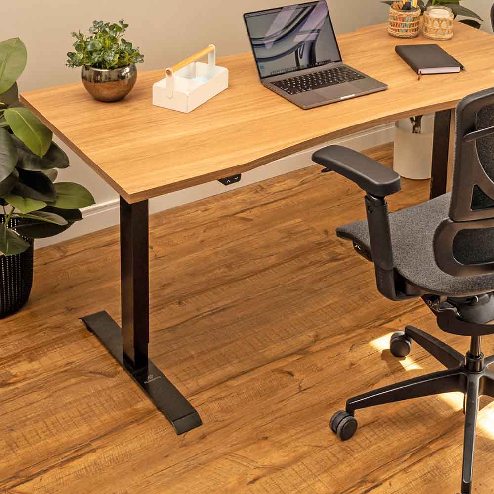 A imagem mostra uma estação de trabalho elegante e acolhedora, com uma cadeira ergonômica de encosto robusto posicionada em frente a uma mesa ajustável em altura Misch com tampo de madeira ergonômico. O ambiente é decorado com plantas e elementos de madeira, além de luminárias pendentes que conferem um toque aconchegante. O piso de madeira e as molduras na parede com arte minimalista completam o espaço, proporcionando um local de trabalho funcional e esteticamente agradável.