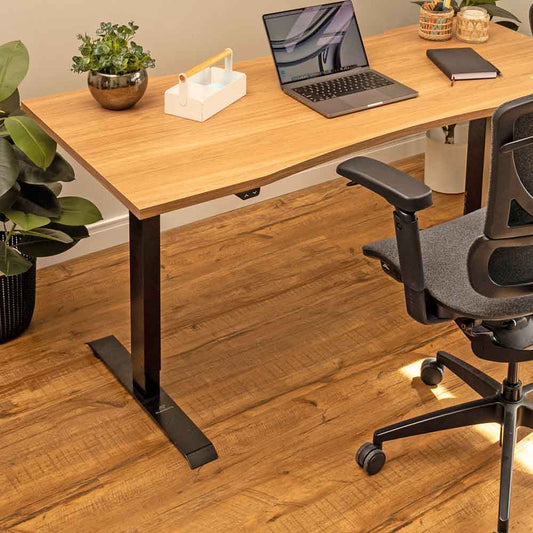 A imagem mostra uma estação de trabalho elegante e acolhedora, com uma cadeira ergonômica de encosto robusto posicionada em frente a uma mesa ajustável em altura Misch com tampo de madeira ergonômico. O ambiente é decorado com plantas e elementos de madeira, além de luminárias pendentes que conferem um toque aconchegante. O piso de madeira e as molduras na parede com arte minimalista completam o espaço, proporcionando um local de trabalho funcional e esteticamente agradável.