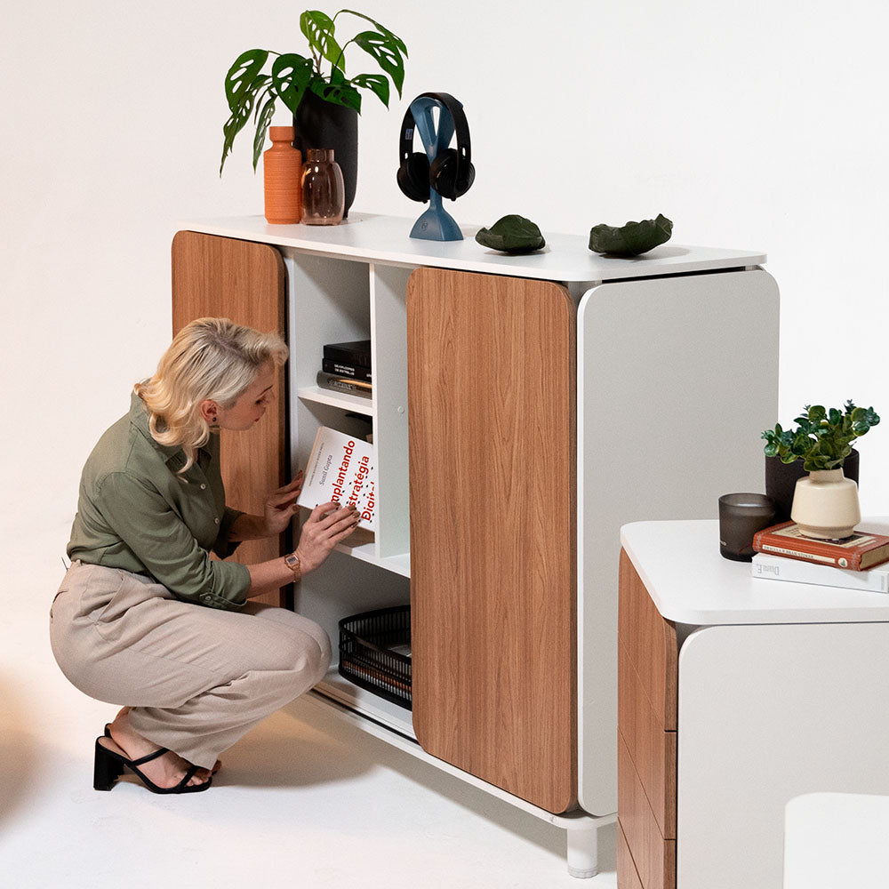 Na imagem se mostra uma moça puxando um livro do armário Appenini Mid na cor Branca, no topo do armário tem vários objetos que combinam com a decoração minimalista. 