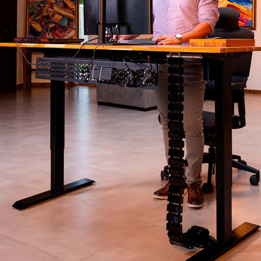 Na imagem, vemos um homem posicionado diante de uma mesa com altura ajustável. Atrás da mesa, encontra-se a calha Spine Cable, que vai da mesa ao chão.